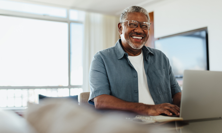 Confident senior man wearing eyeglasses, smiling while using technology on a laptop at home, representing active aging and positivity.